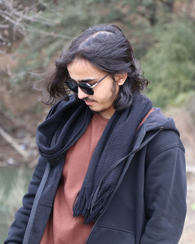 Sparsha outdoors in sunglasses, a black scarf and a rust-coloured sweater under a black hoodie, looking down, with trees behind.