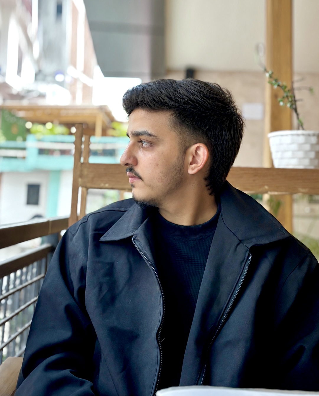 Sparsha in a black jacket over a dark sweater, seated on a wooden balcony and looking off to the side, with a potted plant and buildings behind.